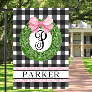May include: A black and white buffalo plaid garden flag with a green wreath and a pink bow. The wreath has a white circle with a black letter "P" in the center. The bottom of the flag has a pink and black patterned band with the name "PARKER" in white.