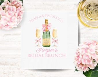 May include: White paper napkin with a watercolor illustration of a champagne bottle, two champagne flutes, and the text "Pearls & Prosecco" and "Harper's Bridal Brunch" in pink script. Pink bows and a crown are also featured.