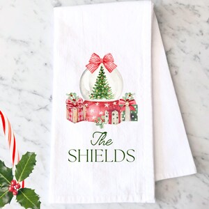 May include: White kitchen towel with a watercolor illustration of a snow globe with a Christmas tree inside, surrounded by presents. The text "The Shields" is printed below the snow globe.