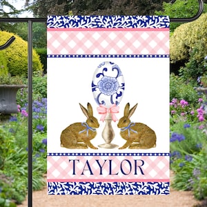 May include: A white garden flag with a blue and white floral egg design, two brown bunnies with blue ribbons, and the name "TAYLOR" in pink letters. The flag has a pink and white gingham pattern at the top and bottom.