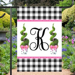 May include: A black and white checkered garden flag with a pink and white polka dot border. The flag features a white background with a black letter K and two topiary trees in white pots with pink bows.