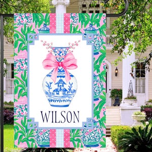 May include: A pink and green garden flag with a blue and white porcelain vase with a pink bow. The vase has a floral design and the text "WILSON" is printed below it.