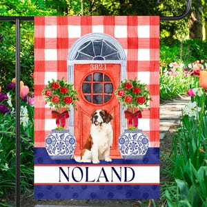 May include: A decorative garden flag featuring a red door with the number 3821, flanked by floral arrangements in blue and white vases. A Saint Bernard dog sits in front of the door, with the name "NOLAND" below. The background has a red and white checkered pattern.