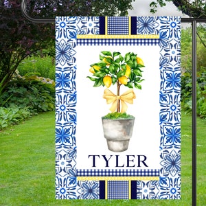 May include: A blue and white garden flag with a lemon tree in a pot. The flag has a yellow bow and the name "TYLER" printed on it.