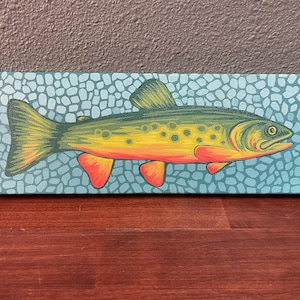 Puede incluir: Una obra de arte rectangular sobre lienzo con una colorida ilustración de un pez. El pez tiene un cuerpo amarillo y verde con aletas naranjas y está sobre un fondo azul con un patrón de guijarros. La obra de arte se exhibe sobre una superficie de madera.