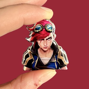 Puede incluir: Un colorido pin de esmalte con un personaje de pelo rojo, gafas azules y una expresión decidida. El personaje lleva una chaqueta azul y negra con detalles dorados. El pin se sujeta entre dos dedos sobre un fondo rojo.