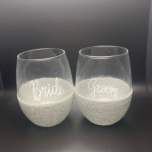 Puede incluir: Dos copas de vino de vidrio transparente con un borde de purpurina plateada. La copa de la izquierda dice "Bride" y la copa de la derecha dice "Groom".