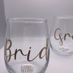 Puede incluir: Dos copas de vino de vidrio transparente con letras doradas. La copa de la izquierda dice "Bride" y "Refill Please". La copa de la derecha dice "Groom".