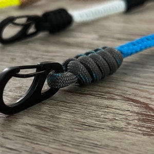 Puede incluir: Un mosquet&oacute;n de metal negro unido a un llavero de paracord gris y azul.