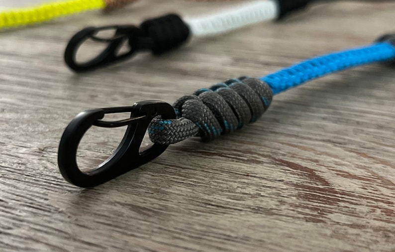 Puede incluir: Un mosquet&oacute;n de metal negro unido a un llavero de paracord gris y azul.
