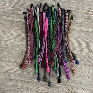 May include: A collection of colorful braided paracord bracelets with various patterns and colors, including green, pink, blue, black, and brown. The bracelets are tied with a knot at the end.