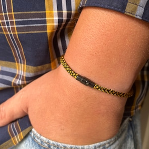 Puede incluir: Una pulsera de cuerda negra y amarilla con un cierre negro se lleva en la muñeca. La pulsera está hecha de un material de cuerda tejida. La persona lleva una camisa a cuadros y vaqueros azules.