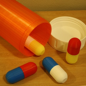 Op de afbeelding: Een oranje plastic pillenfles met een wit deksel en drie kleurrijke capsules. De capsules zijn rood en geel, blauw en wit en blauw en rood.
