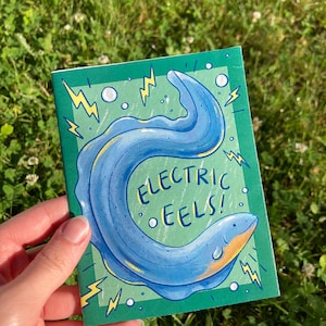 May include: A small, green notebook featuring a blue electric eel illustration with yellow accents. The text "ELECTRIC EELS!" is displayed in a playful font. The notebook is held against a backdrop of green grass.