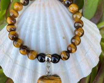 May include: A brown and gold tiger's eye gemstone bracelet with a heart-shaped pendant. The bracelet is made of round beads and has a silver clasp. The pendant is also made of tiger's eye and is attached to the bracelet with a silver chain.
