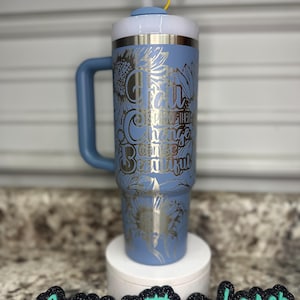 K&ouml;nnte beinhalten: Eine blaue Edelstahl-Reisebecher mit Henkel. Der Becher hat ein silbernes Design mit Sonnenblumen und dem Text "For all the change can be beautiful". Der Becher steht auf einem wei&szlig;en Untersetzer mit einem schwarzen Schild, auf dem in gr&uuml;nen Buchstaben "Nurse with a Laser" steht.
