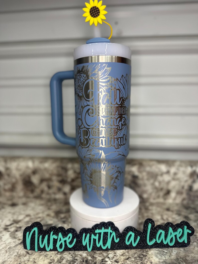 K&ouml;nnte beinhalten: Eine blaue Edelstahl-Reisebecher mit Henkel. Der Becher hat ein silbernes Design mit Sonnenblumen und dem Text "For all the change can be beautiful". Der Becher steht auf einem wei&szlig;en Untersetzer mit einem schwarzen Schild, auf dem in gr&uuml;nen Buchstaben "Nurse with a Laser" steht.