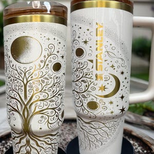 May include: Two white insulated tumblers with gold accents and intricate tree of life and celestial designs. One features the Stanley logo. The text "NURSE WITH A LASER" is printed on the bottom of the tumblers.
