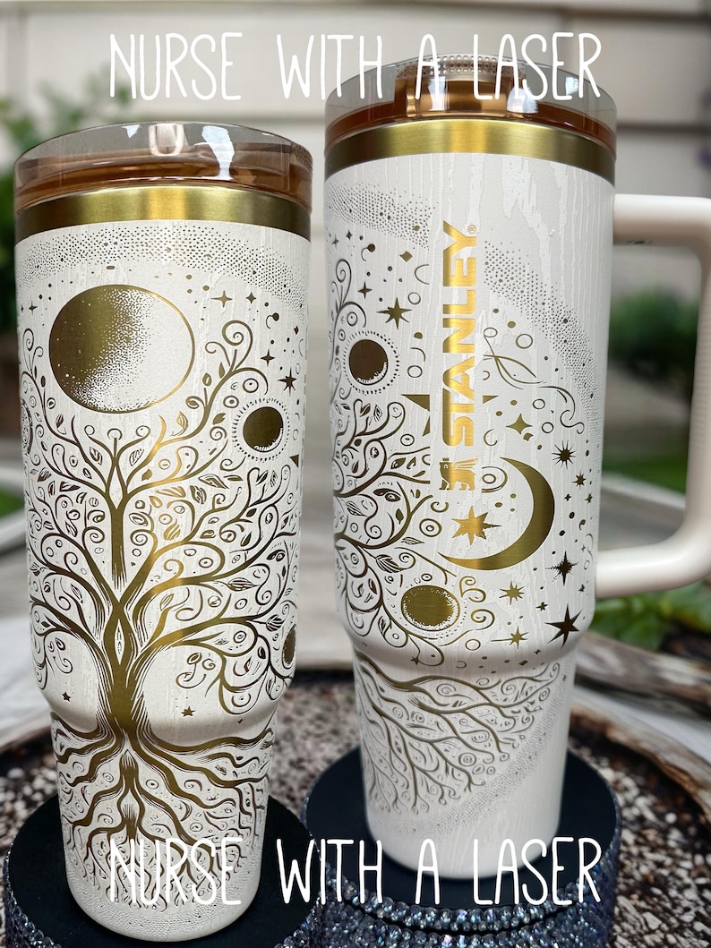 May include: Two white insulated tumblers with gold accents and intricate tree of life and celestial designs. One features the Stanley logo. The text "NURSE WITH A LASER" is printed on the bottom of the tumblers.
