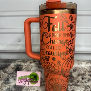 K&ouml;nnte beinhalten: Ein orangefarbener Edelstahlbecher mit Henkel und Strohhalm. Der Becher hat ein goldenes Design aus Bl&auml;ttern, Sonnenblumen und K&uuml;rbissen. Der Text auf dem Becher lautet "Fall is proof that change can be beautiful."