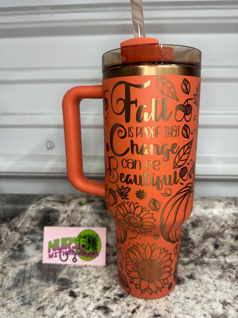 K&ouml;nnte beinhalten: Ein orangefarbener Edelstahlbecher mit Henkel und Strohhalm. Der Becher hat ein goldenes Design aus Bl&auml;ttern, Sonnenblumen und K&uuml;rbissen. Der Text auf dem Becher lautet "Fall is proof that change can be beautiful."