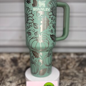 K&ouml;nnte beinhalten: Eine gr&uuml;ne Edelstahl-Reisebecher mit einem schwarzen Griff. Der Becher hat ein Herbst-Design mit K&uuml;rbissen, Bl&auml;ttern und Sonnenblumen. Der Becher ist mit dem Text "STANLEY" graviert.