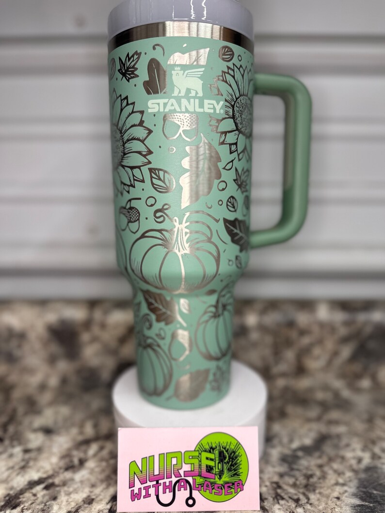 K&ouml;nnte beinhalten: Eine gr&uuml;ne Edelstahl-Reisebecher mit einem schwarzen Griff. Der Becher hat ein Herbst-Design mit K&uuml;rbissen, Bl&auml;ttern und Sonnenblumen. Der Becher ist mit dem Text "STANLEY" graviert.