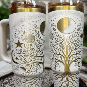 May include: Two white insulated tumblers with gold celestial and tree of life designs. The tumblers have a handle and a clear lid with a gold rim. The text "NURSE WITH A LASER" is printed on the top and bottom of the tumblers.
