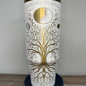 May include: A white tumbler with a gold lid and a gold design. The design features a tree of life with a moon and stars. The tumbler is approximately 9 inches tall.