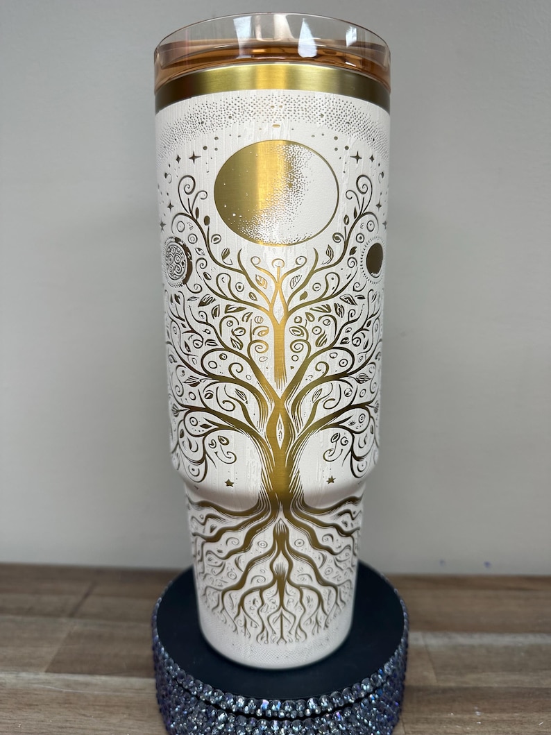 May include: A white tumbler with a gold lid and a gold design. The design features a tree of life with a moon and stars. The tumbler is approximately 9 inches tall.