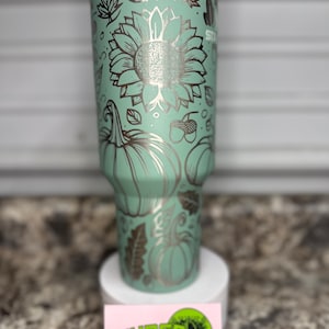 K&ouml;nnte beinhalten: Ein Edelstahlbecher mit einer hellgr&uuml;nen Pulverbeschichtung. Der Becher ist mit einem Laser-gravierten Design aus K&uuml;rbissen, Sonnenblumen, Bl&auml;ttern und Eicheln verziert. Der Becher steht auf einer wei&szlig;en Oberfl&auml;che mit einer pinkfarbenen und gr&uuml;nen Visitenkarte, auf der "Nurse with a Laser" steht.