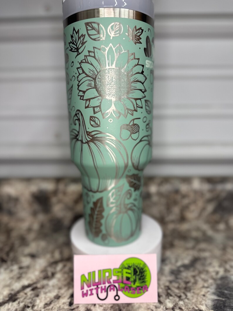 K&ouml;nnte beinhalten: Ein Edelstahlbecher mit einer hellgr&uuml;nen Pulverbeschichtung. Der Becher ist mit einem Laser-gravierten Design aus K&uuml;rbissen, Sonnenblumen, Bl&auml;ttern und Eicheln verziert. Der Becher steht auf einer wei&szlig;en Oberfl&auml;che mit einer pinkfarbenen und gr&uuml;nen Visitenkarte, auf der "Nurse with a Laser" steht.