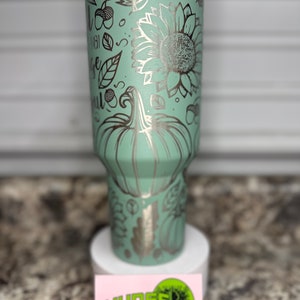 K&ouml;nnte beinhalten: Ein hellgr&uuml;ner Edelstahlbecher mit einem silbernen Design aus K&uuml;rbissen, Sonnenblumen, Bl&auml;ttern und Eicheln. Der Becher hat einen wei&szlig;en Deckel und steht auf einer wei&szlig;en Oberfl&auml;che.