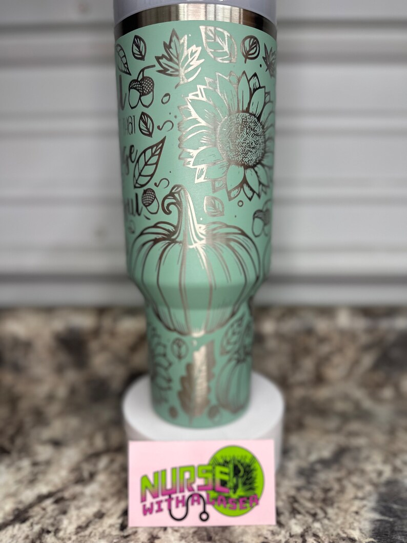 K&ouml;nnte beinhalten: Ein hellgr&uuml;ner Edelstahlbecher mit einem silbernen Design aus K&uuml;rbissen, Sonnenblumen, Bl&auml;ttern und Eicheln. Der Becher hat einen wei&szlig;en Deckel und steht auf einer wei&szlig;en Oberfl&auml;che.