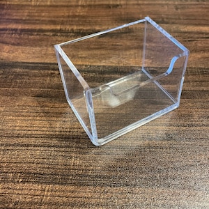 Puede incluir: Un tarjetero rectangular transparente de acr&iacute;lico con un recorte curvo en un lado. El soporte est&aacute; abierto en la parte superior e inclinado, dise&ntilde;ado para mostrar tarjetas. El soporte est&aacute; sobre una superficie de madera.