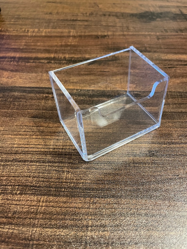 Puede incluir: Un tarjetero rectangular transparente de acr&iacute;lico con un recorte curvo en un lado. El soporte est&aacute; abierto en la parte superior e inclinado, dise&ntilde;ado para mostrar tarjetas. El soporte est&aacute; sobre una superficie de madera.