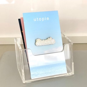 Puede incluir: Un tarjetero de acr&iacute;lico transparente con una pila de tarjetas. La tarjeta superior es de color azul claro con la palabra "utopie" y un pin "Breathe". Se ven otras tarjetas en el soporte. El soporte est&aacute; sobre una superficie gris.