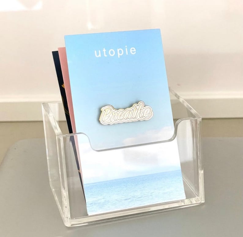 Puede incluir: Un tarjetero de acr&iacute;lico transparente con una pila de tarjetas. La tarjeta superior es de color azul claro con la palabra "utopie" y un pin "Breathe". Se ven otras tarjetas en el soporte. El soporte est&aacute; sobre una superficie gris.
