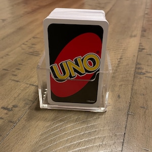 Puede incluir: Un mazo de cartas UNO en un soporte de acr&iacute;lico transparente. El reverso de las cartas presenta el logotipo de UNO en blanco y amarillo sobre un &oacute;valo rojo sobre un fondo negro. El soporte es rectangular y est&aacute; sobre una superficie de madera.
