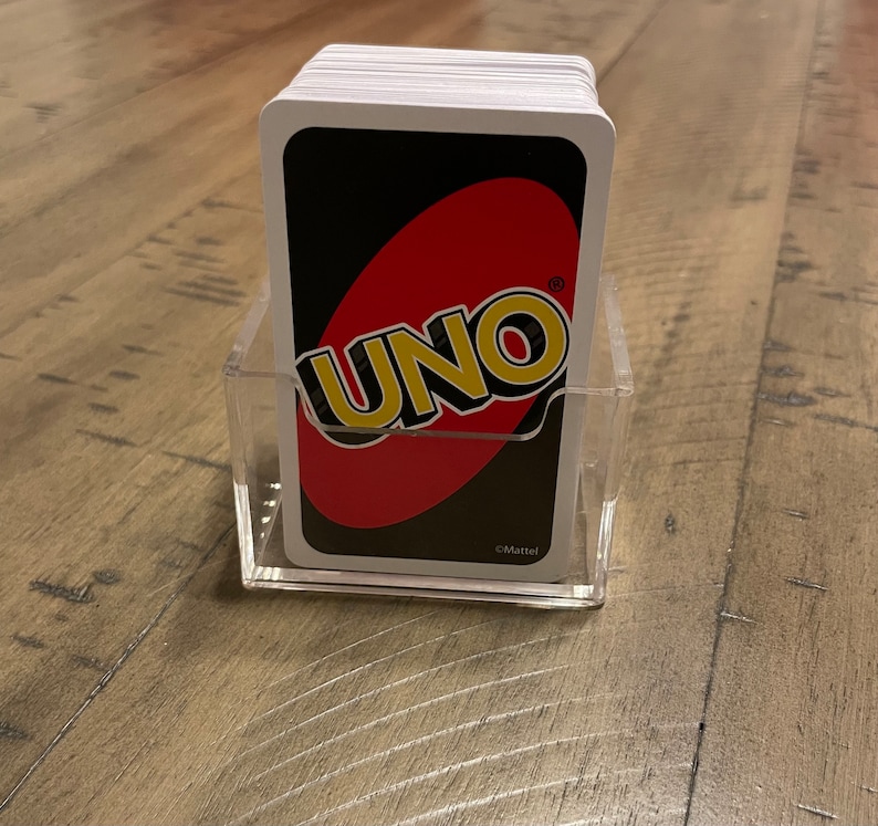 Puede incluir: Un mazo de cartas UNO en un soporte de acr&iacute;lico transparente. El reverso de las cartas presenta el logotipo de UNO en blanco y amarillo sobre un &oacute;valo rojo sobre un fondo negro. El soporte es rectangular y est&aacute; sobre una superficie de madera.