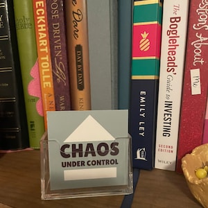 Puede incluir: Un tarjetero de acr&iacute;lico transparente con una tarjeta azul claro que dice "CHAOS UNDER CONTROL". La tarjeta tiene un dise&ntilde;o de tri&aacute;ngulo blanco en la parte superior. Libros de varios colores y t&iacute;tulos en el fondo.