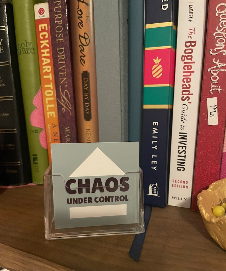 Puede incluir: Un tarjetero de acr&iacute;lico transparente con una tarjeta azul claro que dice "CHAOS UNDER CONTROL". La tarjeta tiene un dise&ntilde;o de tri&aacute;ngulo blanco en la parte superior. Libros de varios colores y t&iacute;tulos en el fondo.