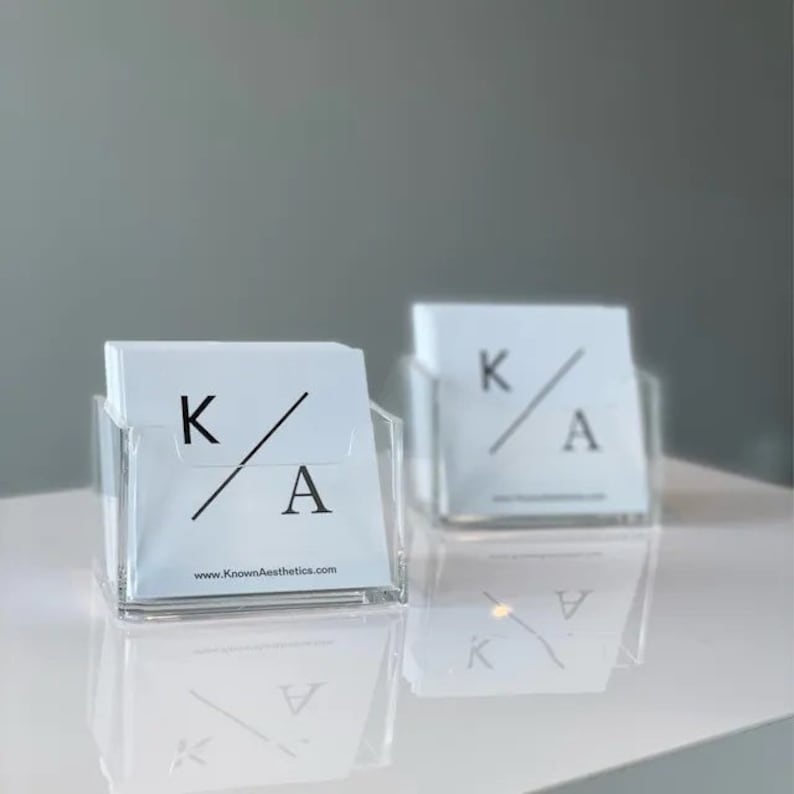 Puede incluir: Dos tarjeteros transparentes de acr&iacute;lico con tarjetas blancas. Cada tarjeta muestra las letras "K" y "A" con una l&iacute;nea diagonal y la direcci&oacute;n del sitio web "KnownAesthetics.com". Los tarjeteros est&aacute;n sobre una superficie blanca.