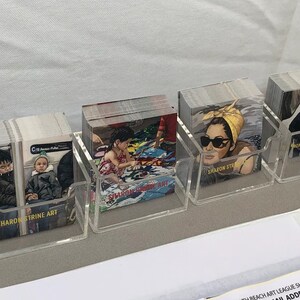 Puede incluir: Varias pilas de tarjetas de arte exhibidas en soportes de acr&iacute;lico transparente. Las tarjetas presentan diversas im&aacute;genes, como un ni&ntilde;o en la playa, una mujer con gafas de sol y personas en el metro. El nombre del artista, "SHARON STRINE ART", es visible.