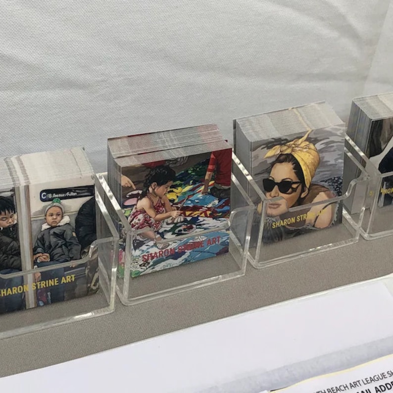 Puede incluir: Varias pilas de tarjetas de arte exhibidas en soportes de acr&iacute;lico transparente. Las tarjetas presentan diversas im&aacute;genes, como un ni&ntilde;o en la playa, una mujer con gafas de sol y personas en el metro. El nombre del artista, "SHARON STRINE ART", es visible.