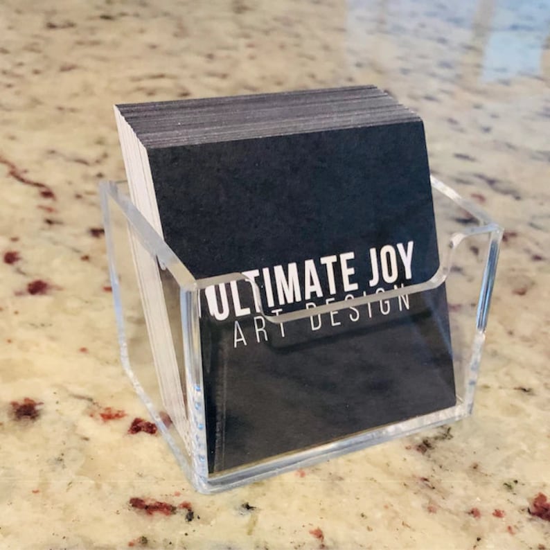 Puede incluir: Un tarjetero de acr&iacute;lico transparente lleno de tarjetas de visita negras. Las tarjetas muestran el texto "ULTIMATE JOY ART DESIGN" en blanco. El soporte se encuentra sobre una superficie clara y moteada.