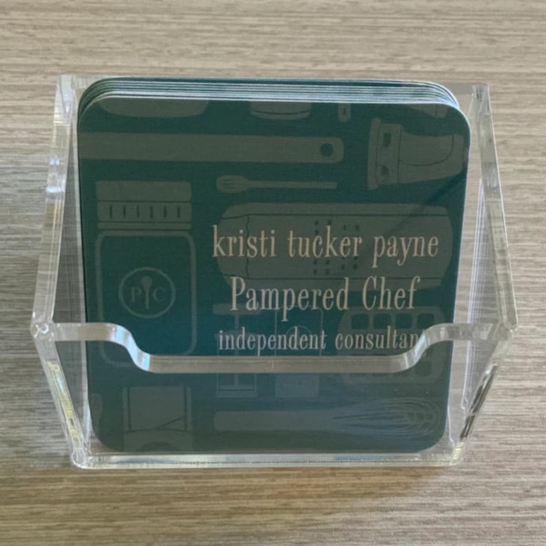 Puede incluir: Un tarjetero de acr&iacute;lico transparente que contiene varias tarjetas de visita de color verde azulado. Las tarjetas presentan ilustraciones de utensilios de cocina y el texto "kristi tucker payne Pampered Chef independent consultant."