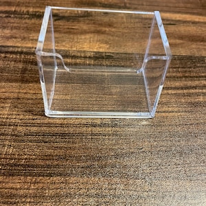 Puede incluir: Un tarjetero transparente de acr&iacute;lico, con base rectangular y lados angulados. El soporte est&aacute; dise&ntilde;ado para mostrar las tarjetas en posici&oacute;n vertical y est&aacute; colocado sobre una superficie de madera.