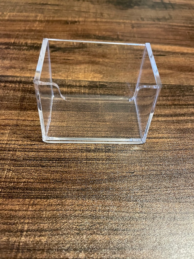 Puede incluir: Un tarjetero transparente de acr&iacute;lico, con base rectangular y lados angulados. El soporte est&aacute; dise&ntilde;ado para mostrar las tarjetas en posici&oacute;n vertical y est&aacute; colocado sobre una superficie de madera.