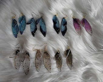 May include: A set of eight pairs of earrings featuring delicate, translucent wings with gold accents. The wings are in shades of blue, purple, and black.